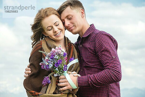 Mädchen in einem karierten und mit einem Blumenstrauß umarmt mit ihrem Geliebten auf einem Hintergrund von einem sanft blaün Himmel