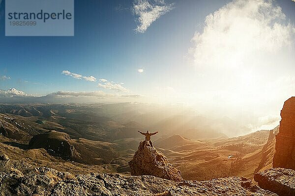 Ein glücklicher Mann mit hoch erhobenen Händen steht auf einem separat stehenden Felsen  der sich über den Wolken befindet  vor dem Hintergrund von Tälern  Hügeln  Sonnenuntergang und einem schönen Himmel