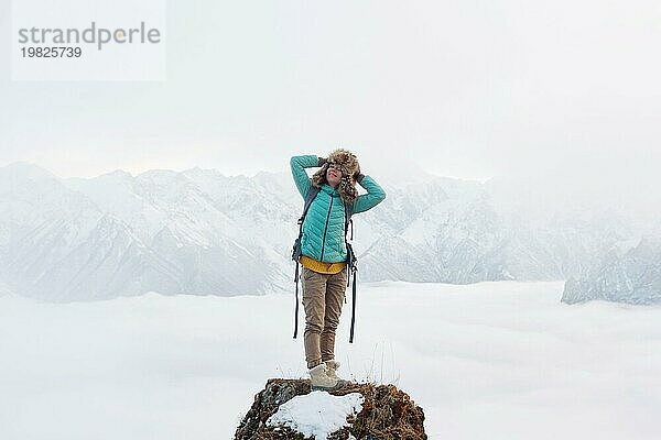 Ein fröhlich lächelndes Hipster Mädchen reist in einer Daunenjacke mit einem Rucksack und einem großen Pelzhut und steht auf einem Felsen vor dem Hintergrund verschwommener Berge und einem Tal in den Wolken unter ihr. Das Konzept des Reisens