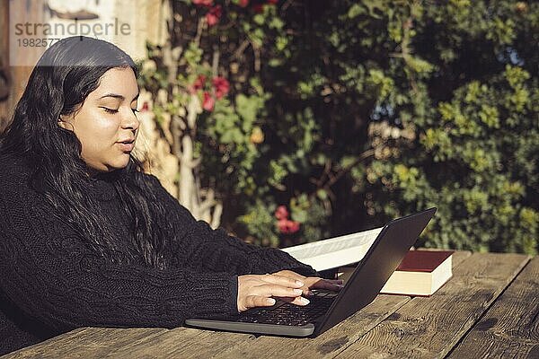 Lateinamerikanische Frau mit langen dunklen Haaren  die an ihrem Laptop tippt und an einem universitären Bildungsprojekt arbeitet  draußen im Park auf dem Universitätscampus