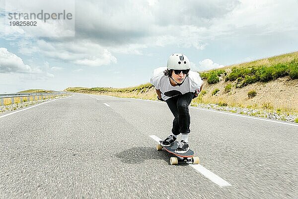 Der Typ mit dem Helm und der Sonnenbrille fährt mit seinem Longboard in einem speziellen Sportständer sehr schnell über die asphaltierte Straße