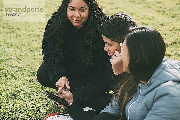 Eine hispanische Familie verbringt an einem schönen  sonnigen Tag Zeit miteinander in einem örtlichen Park. Ein Teenager sitzt im Gras  hält sein Smartphone in der Hand und schaut weg. Seine Mutter und seine Schwester sitzen neben ihm  schauen in die Kamera und lächeln