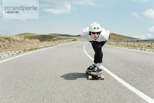 Der Typ mit dem Helm und der Sonnenbrille fährt mit seinem Longboard in einem speziellen Sportständer sehr schnell über die asphaltierte Straße