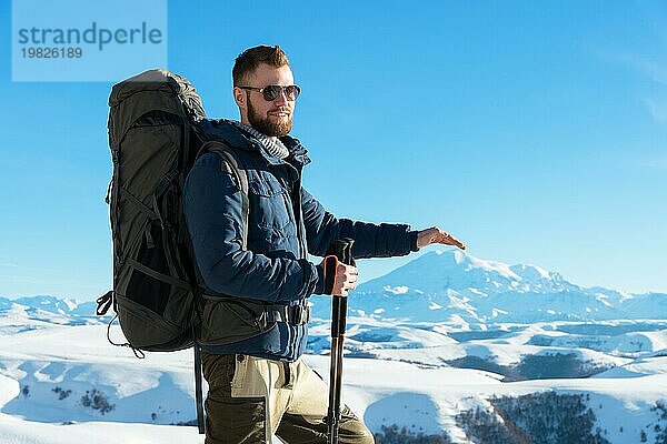 Ein Hipster Reisender mit Bart und Sonnenbrille in der Natur. Ein Mann wandern in den Bergen mit einem Rucksack und skandinavischen Wanderstöcke im Hintergrund eines Berges Elbrus Landschaft und blaün Himmel. Reisen Lifestyle Abenteuer Outdoor Erholung übertreffen