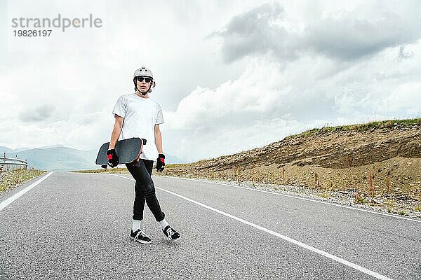 Ein junger Mann  ein Skater mit Helm  Brille und Handschuhen  wartet mit einem Longboard in der Hand auf ein Rennen auf einer Landstraße