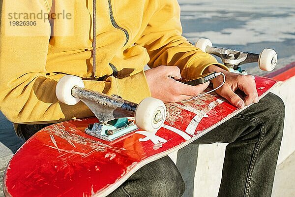 Nahaufnahme eines Teenagers in Sweatshirt  Jeans und Turnschuhen  der in einem Skatepark sitzt und ein Telefon mit Kopfhörern und ein Skateboard hält