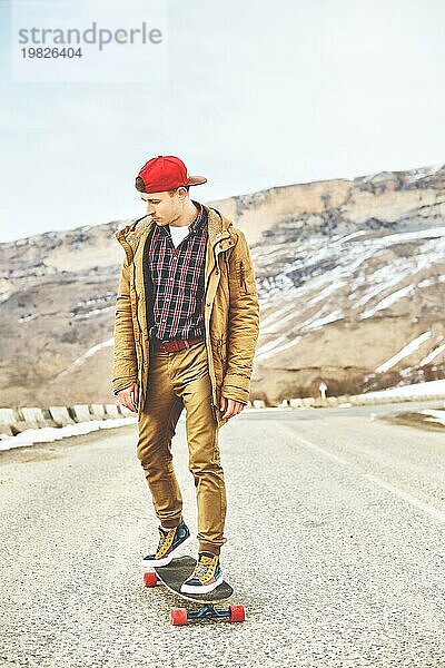 Stylish happy Junger Mann in Mütze und Hose Jogger rollen auf einem Longboard eine Bergstraße hinunter  genießen das Leben