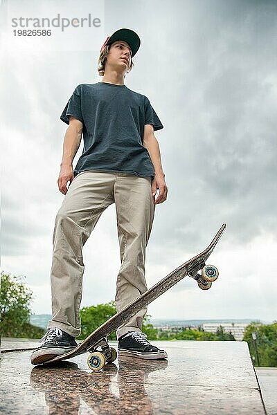 Ein junger langhaariger Skater im TShirt und mit einer langen Mütze steht mit einem Skateboard vor dem Hintergrund einer Stadtlandschaft