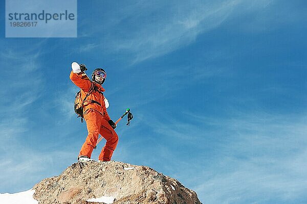 Porträt eines Skifahrers in einem orangefarbenen Overall mit einem Rucksack auf dem Rücken und Skiern auf seinen Schultern in einem Helm steht auf einem Felsen gegen den blaün Himmel. Kopieren Sie den Raum. Das Konzept der aktiven Erholung im Winter und Skifahren