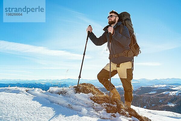 Ein Hipster Reisender mit Bart und Sonnenbrille in der Natur. Ein Mann wandert in den Bergen mit einem Rucksack und skandinavischen Wanderstöcken vor dem Hintergrund einer Berglandschaft und blauem Himmel. Reisen Lifestyle Abenteuer Outdoor Erholung Outdoor Sport