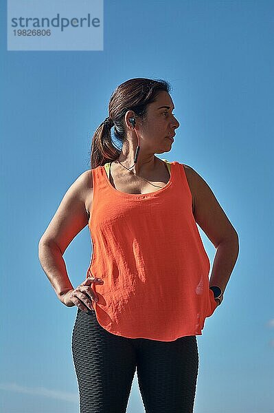 Eine Frau in Fitnesskleidung steht selbstbewusst vor einem klaren blauen Himmel