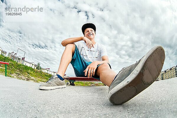 Ein junger lachender Skater in Mütze und Shorts sitzt auf seinem Skateboard in einem Skatepark