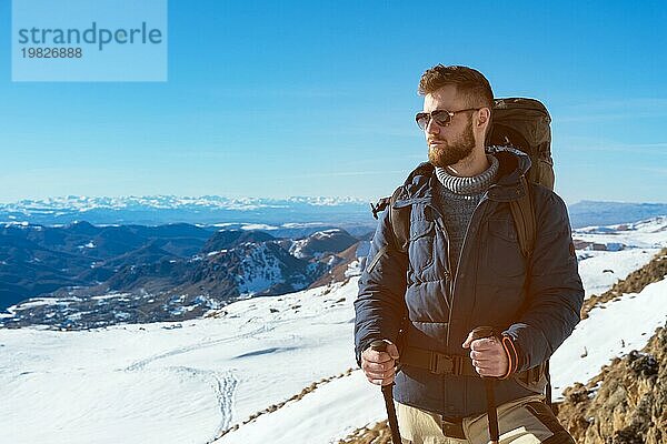 Ein Hipster Reisender mit Bart und Sonnenbrille in der Natur. Ein Mann wandert in den Bergen mit einem Rucksack und skandinavischen Wanderstöcken vor dem Hintergrund einer Berglandschaft und blauem Himmel. Reisen Lifestyle Abenteuer Outdoor Erholung Outdoor Sport