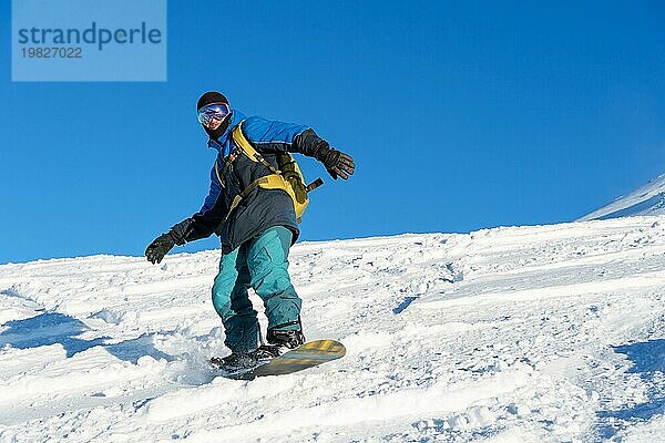 Ein Freeboard Snowboarder mit Skimaske und Rucksack fährt über die schneebedeckte Piste und hinterlässt einen Pulverschnee vor dem blaün Himmel