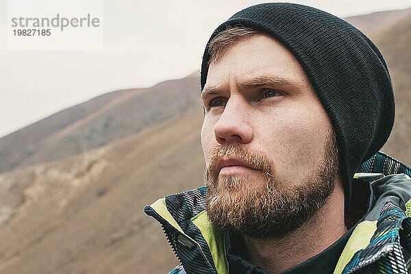 Ein strenger Hipster Typ mit Bart und Blick in den Hut in der Ferne in den Bergen  ein großes Porträt