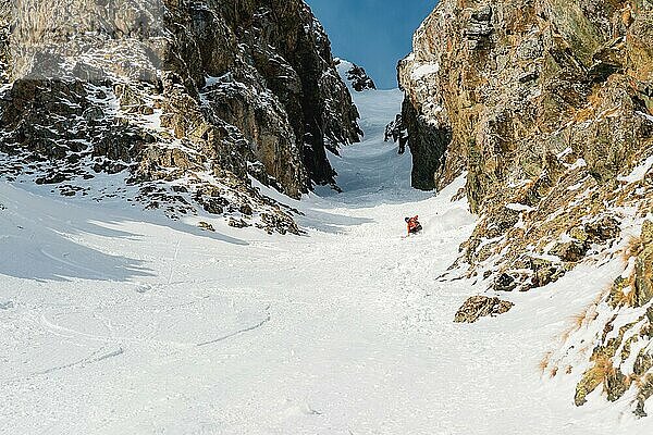 Ein männlicher Freerider mit Bart fährt im Backcountry mit hoher Geschwindigkeit von der Piste ab und hinterlässt vor dem Hintergrund epischer Felsen eine Spur aus Pulverschnee. Das Konzept der Freeride Kultur und Backcountry Destinationen im Extremskisport