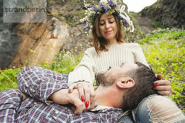 Hipster mit Bart liegt auf dem Schoß seines Mädchens  das ein Wildblumen Vinok trägt. Das Konzept der Einheit mit der Natur und ein junges Paar von Menschen