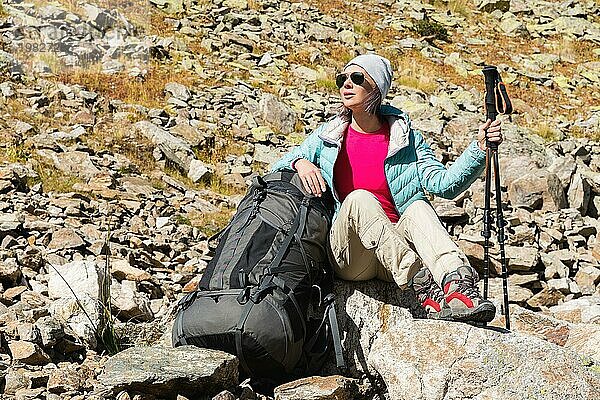 Ein Touristenmädchen mit Sonnenbrille  Daunenjacke und Hut  mit einem Rucksack und einer Bergausrüstung mit Griffen zum Wandern in den Händen  sitzt und entspannt sich und genießt die schöne Aussicht auf einen Hochgebirgssee im Nordkaukasus. Karatschai Tscherkess R