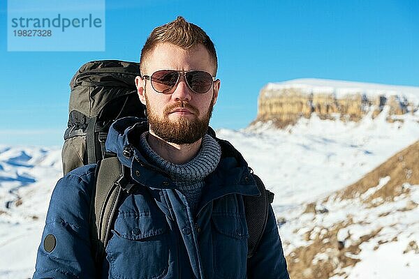 Ein Hipster Reisender mit Bart und Sonnenbrille in der Natur. Ein Mann wandert in den Bergen mit einem Rucksack und skandinavischen Wanderstöcken vor dem Hintergrund einer Berglandschaft und blauem Himmel. Reisen Lifestyle Abenteuer Outdoor Erholung Outdoor Sport