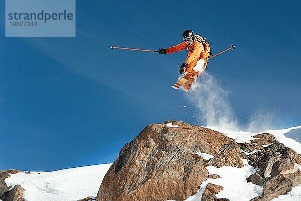 Ein professioneller Skifahrer springt von einer hohen Klippe gegen den blaün Himmel und hinterlässt eine Spur aus Pulverschnee in den Bergen. Foto von den Hängen des Berges Elbrus. Das Konzept des Extremsports und der Erholung in den Bergen im Winter. Kopieren Sie den Raum