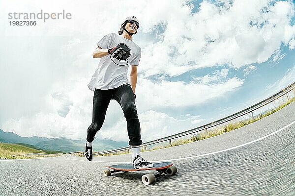 Ein Mann mit Helm und Sonnenbrille beschleunigt mit dem Fuß auf seinem Longboard auf dem Asphalt einer Landstraße und lächelt. Weitwinkel