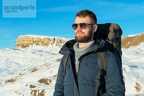 Ein Hipster Reisender mit Bart und Sonnenbrille in der Natur. Ein Mann wandern in den Bergen mit einem Rucksack und skandinavischen Wanderstöcke im Hintergrund eines Berges Elbrus Landschaft und blaün Himmel. Reisen Lifestyle Abenteuer Outdoor Erholung übertreffen