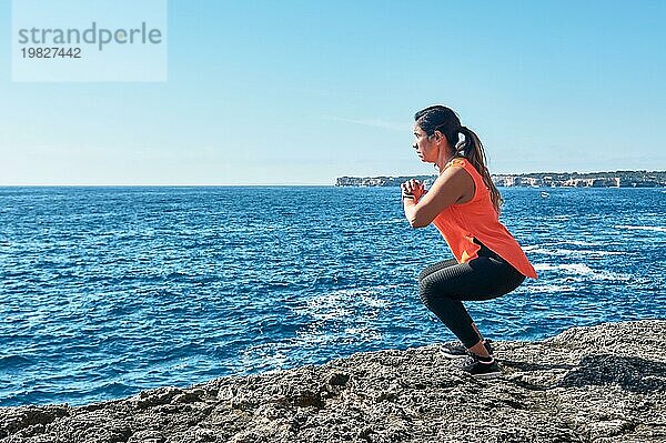 Eine Frau hockt an einem sonnigen Tag am Meer  gekleidet in Trainingskleidung  mit heiterer Miene