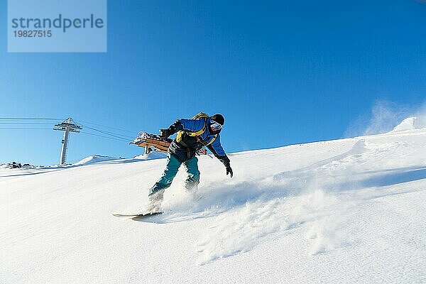 Ein Freeboard Snowboarder mit Skimaske und Rucksack fährt über die schneebedeckte Piste und hinterlässt einen Pulverschnee vor dem blaün Himmel