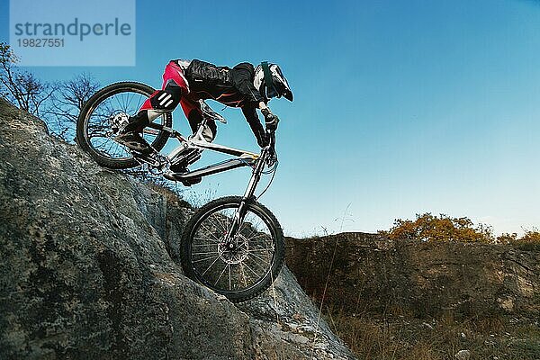 Ein junger Fahrer auf einem Mountainbike kommt von einer Klippe vor einem blaün Himmel herunter. Downhill Richtung im Radsport ist eine Geschwindigkeit bergab vom Berg auf einem Fahrrad. Konzept des extrem und Enduro Radsports
