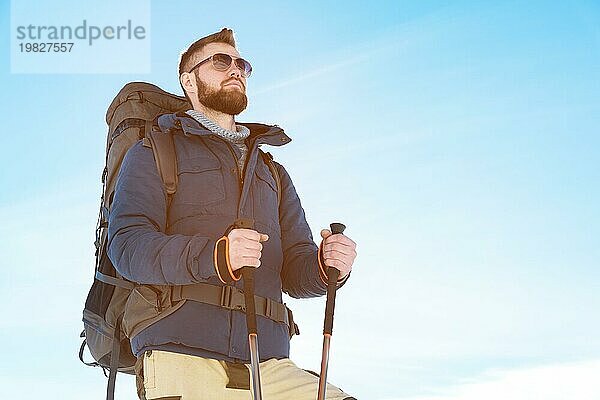 Ein Hipster Reisender mit Bart und Sonnenbrille in der Natur. Ein Mann wandert in den Bergen mit einem Rucksack und skandinavischen Wanderstöcken vor dem Hintergrund einer Berglandschaft und blauem Himmel. Reisen Lifestyle Abenteuer Outdoor Erholung Outdoor Sport