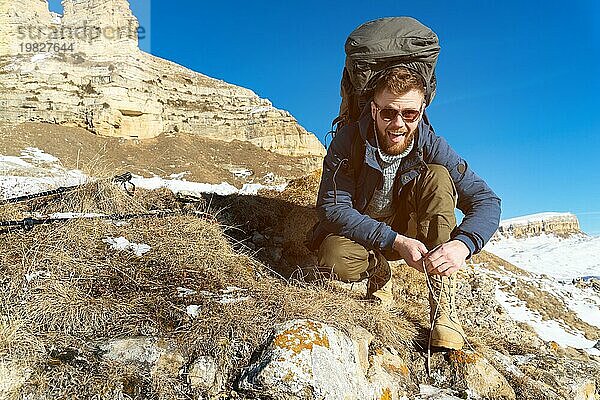 Porträt eines glücklich lachenden Hipster Reisenden mit Bart und Sonnenbrille sitzt und bindet Schnürsenkel in der Natur. Ein Mann wandert in den Bergen mit einem Rucksack und skandinavischen Wanderstöcken im Hintergrund einer Berglandschaft. Reisen Leben