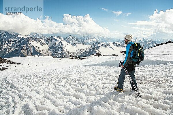 Müder Reisender mit Sonnenbrille und einem Rucksack  auf dem der Eispickel von einem schneebedeckten Gipfel herabhängt  vor dem Hintergrund schneebedeckter Berge und mit einem Stock für skandinavisches Wandern