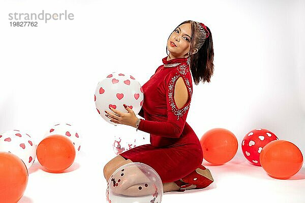 Junge sinnliche Frau mit rotem Kleid schaut in die Kamera und spielt mit Luftballons. Studio Porträt auf weißem Hintergrund. Konzept der Selbstliebe