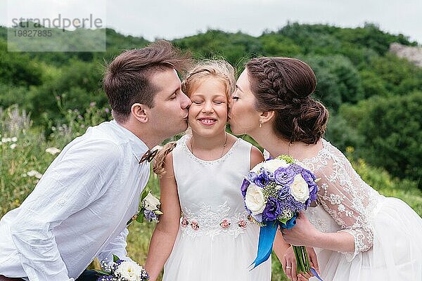 Junge Eltern auf einem Hochzeitsspaziergang in Hochzeitskleidern küssen ihre junge Tochter in die Wangen. Konzept einer glücklichen jungen Familie