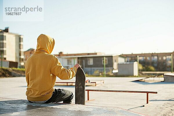 Nahaufnahme eines jungen Hipsters in Sweatshirt und Jeans  der traurig in einem Skatepark sitzt und ein Skateboard hält