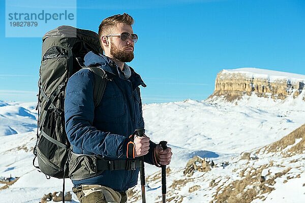 Ein Hipster Reisender mit Bart und Sonnenbrille in der Natur. Ein Mann wandert in den Bergen mit einem Rucksack und skandinavischen Wanderstöcken vor dem Hintergrund einer Berglandschaft und blauem Himmel. Reisen Lifestyle Abenteuer Outdoor Erholung Outdoor Sport