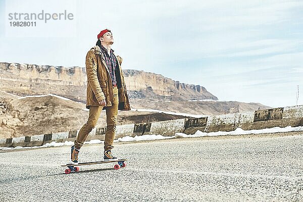 Stylish happy Junger Mann in Mütze und Hose Jogger rollen auf einem Longboard eine Bergstraße hinunter  genießen das Leben