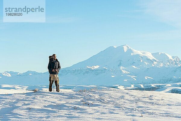 Ein Wanderer mit einem großen Rucksack auf den Schultern steht auf einem schneebedeckten Hügel vor dem blaün Himmel und dem schlafenden Vulkan Elbrus  der vom kaukasischen Hauptkamm umgeben ist  und meditiert. Das Konzept des Wanderns und der Harmonie durch Meditation in der Natur in win