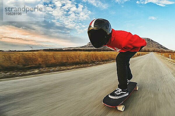 Ein junger Mann in einem Fullface Helm und einem Lederanzug in einem speziellen Gestell fährt ein Longboard auf afsaltu Sonnenuntergang im Hintergrund Berge und schönen Himmel mit hoher Geschwindigkeit