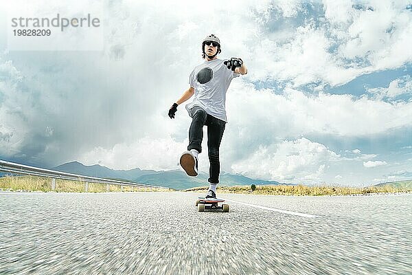 Ein Mann mit Helm und Sonnenbrille beschleunigt mit dem Fuß auf seinem Longboard auf dem Asphalt einer Landstraße und lächelt. Weitwinkel