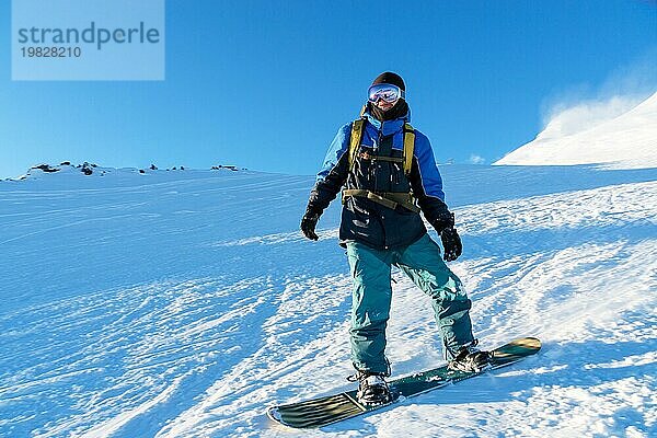 Ein Freeboard Snowboarder mit Skimaske und Rucksack fährt über die schneebedeckte Piste und hinterlässt einen Pulverschnee vor dem blaün Himmel