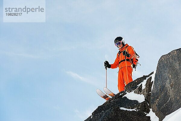 Ein professioneller Skisportler steht an einem sonnigen Tag am Rande eines hohen Felsens vor dem blaün Himmel. Kopieren Sie den Raum. Das Konzept des Skifahrens