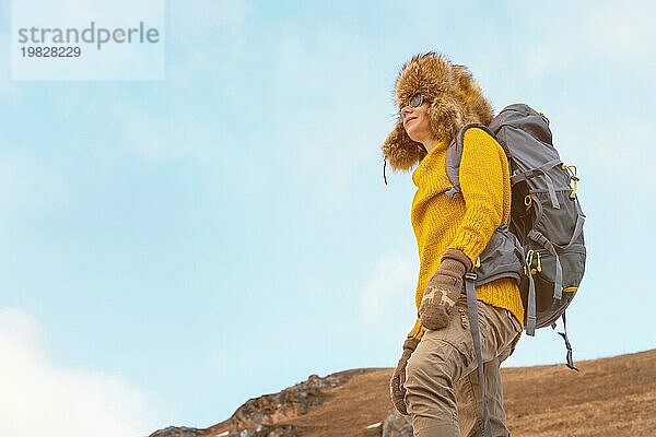 Das Backpacker Mädchen mit Sonnenbrille und einer großen nordischen Pelzmütze  das einen Rucksack auf dem Rücken trägt  steht auf einem Felsen und blickt in Richtung der Klippen  die sich im Felsen verstecken. Das Konzept der Erholung in den Bergen und des Tourismus. Urlaub in den Bergen. Ansicht von hinten