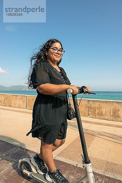 Eine fröhliche Frau fährt an einem sonnigen Tag mit einem Elektroroller am Meer entlang