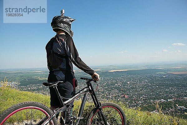 Ein junger Kerl steht auf Ihrem Mountainbike auf einem Berg  wenn unterhalb der Berge niedrige Wolken und Nebel