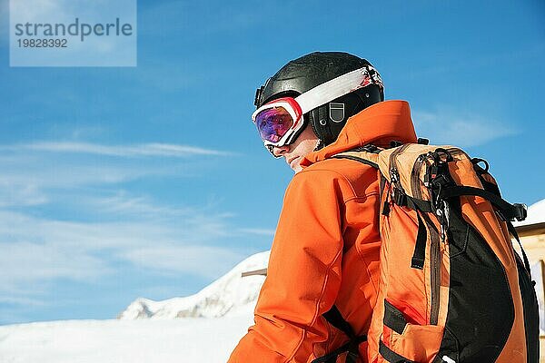 Porträt eines Skifahrers in einem orangefarbenen Overall mit einem Rucksack auf dem Rücken in einem Helm steht vor dem Hintergrund einer schönen kaukasischen Berglandschaft mit schneebedeckten Bergen des kaukasischen Kammes und blaün Himmel. Kopieren Sie das Konzept der aktiven recreati