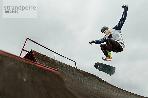 Teenager Skateboarder in einer Kappe macht einen Tricksprung auf einer Halfpipe vor einem bewölkten Himmel