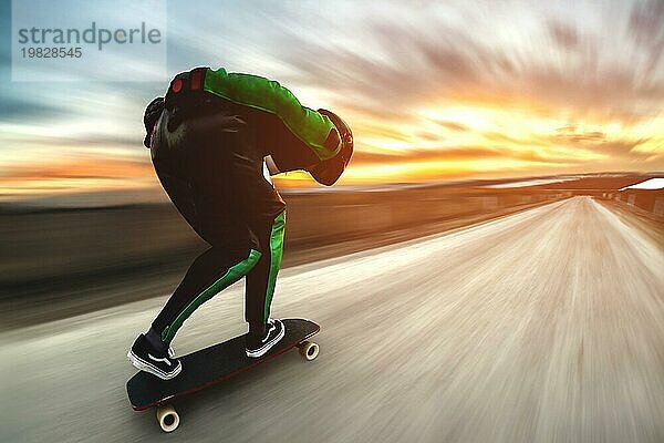 Ein Mann in Helm und Lederkombi fährt in einem speziellen Gestell mit hoher Geschwindigkeit auf einem Longboard bergab auf Afsalt vor der Kulisse eines schönen Sonnenuntergangshimmels im Gegenlicht der untergehenden Sonne. Der Hintergrund ist dynamisch aufgefächert