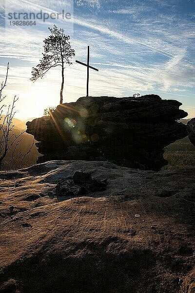 Landschaftsaufnahme auf einem Sandsteinfelsen im Wald. Morgenstimmung bei Sonnenaufgang an einem Aussichtspunkt. Ein kleiner Baum und ein Gipfelkreuz stehen auf dem Rötzenfels im Pfälzerwald