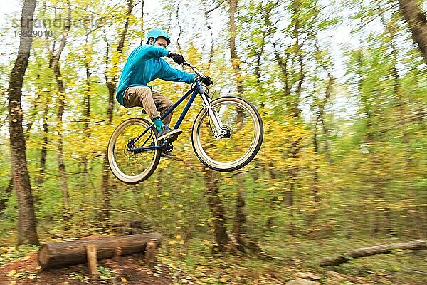 Ein junger Fahrer mit Helm und blauem Sweatshirt fliegt auf einem Fahrrad  nachdem er von einem hohen Kicker auf einem Waldradweg gesprungen ist. Aufnahme mit Langzeitbelichtung Verdrahtung